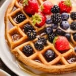 A golden waffle topped with strawberries, blackberries, blueberries, cream, and syrup sits on a white plate.