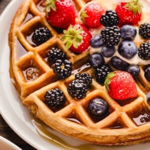A golden waffle topped with strawberries, blackberries, blueberries, cream, and syrup sits on a white plate.