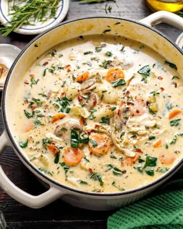 A creamy soup with shredded chicken, carrots, spinach, and herbs in a white pot on a dark table, surrounded by garlic, herbs, and a green towel.