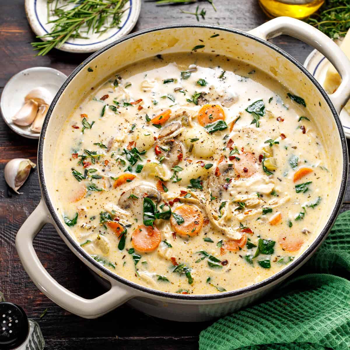 A creamy soup with shredded chicken, carrots, spinach, and herbs in a white pot on a dark table, surrounded by garlic, herbs, and a green towel.