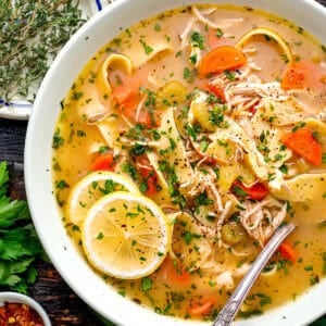 A bowl of chicken noodle soup with carrots, celery, herbs, and lemon slices, garnished with parsley.