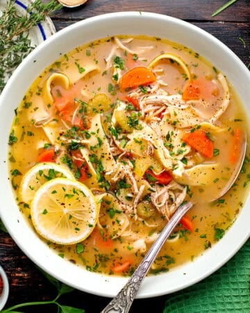 A bowl of chicken noodle soup topped with fresh herbs and lemon slices, with chunks of chicken, carrots, celery, and noodles in a clear broth.