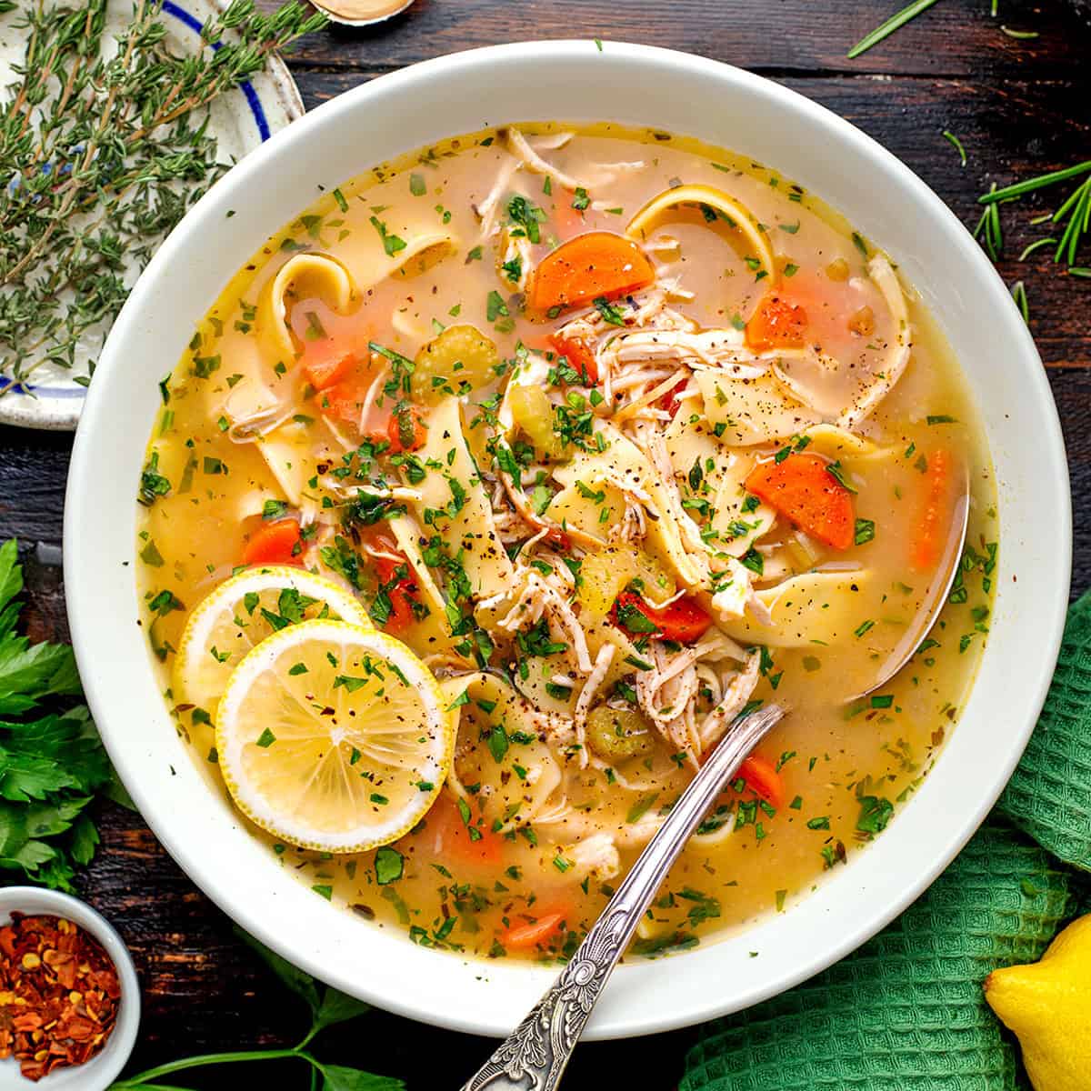 A bowl of chicken noodle soup topped with fresh herbs and lemon slices, with chunks of chicken, carrots, celery, and noodles in a clear broth.