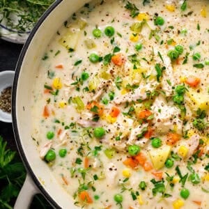 Creamy chicken and vegetable soup with peas, carrots, corn, potatoes, and herbs in a white pot, garnished with black pepper and fresh parsley.