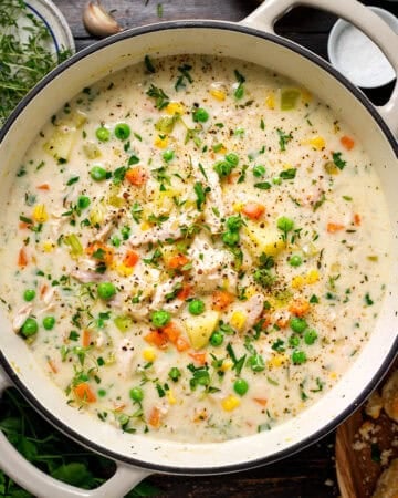 A creamy vegetable and chicken soup in a large white pot, topped with fresh herbs, peas, carrots, and corn.
