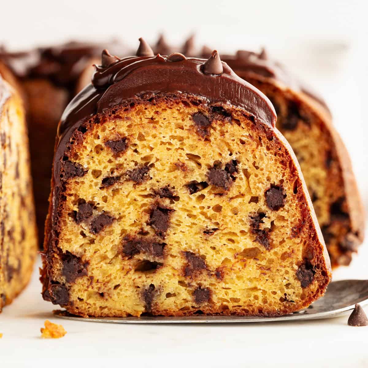 A close-up of a chocolate chip bundt cake slice topped with chocolate glaze and chocolate chips, showing a moist, golden interior with plenty of chocolate chips.