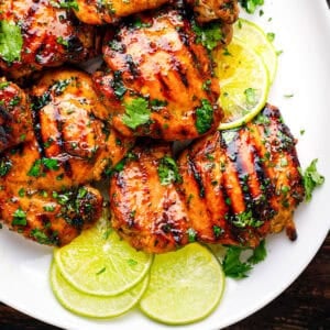 Grilled chicken thighs glazed with sauce, garnished with chopped cilantro, and served on a white plate with fresh lime slices.