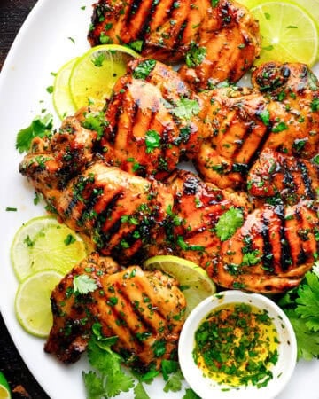 Grilled chicken thighs garnished with fresh cilantro and lime slices on a white plate, with a small bowl of herb oil and additional limes on the side.