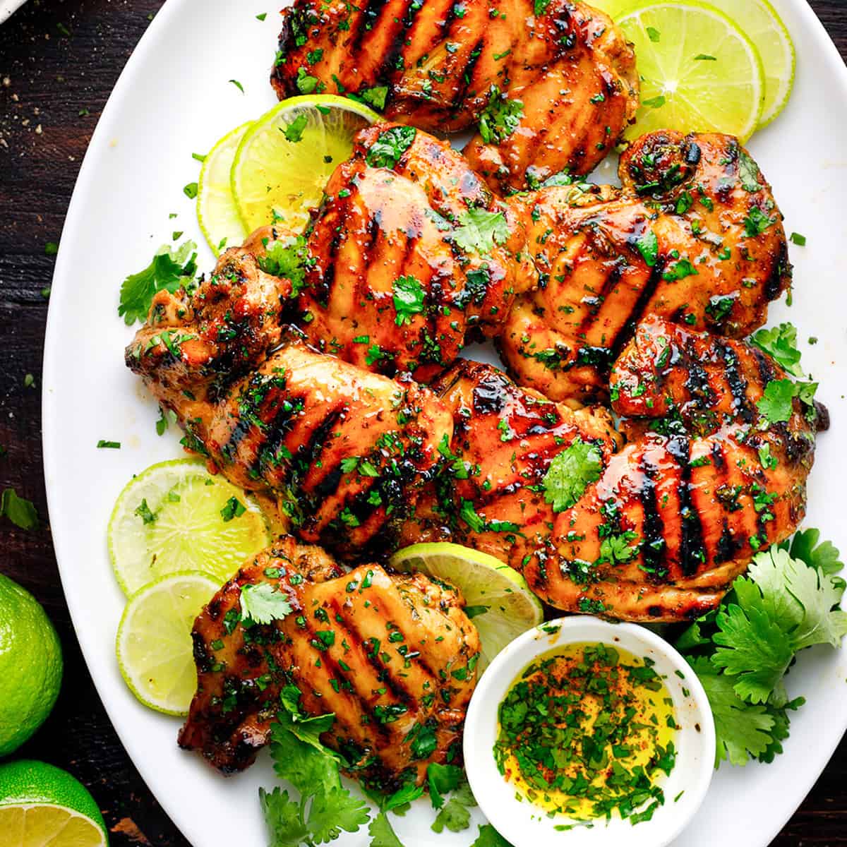 Grilled chicken thighs garnished with fresh cilantro and lime slices on a white plate, with a small bowl of herb oil and additional limes on the side.