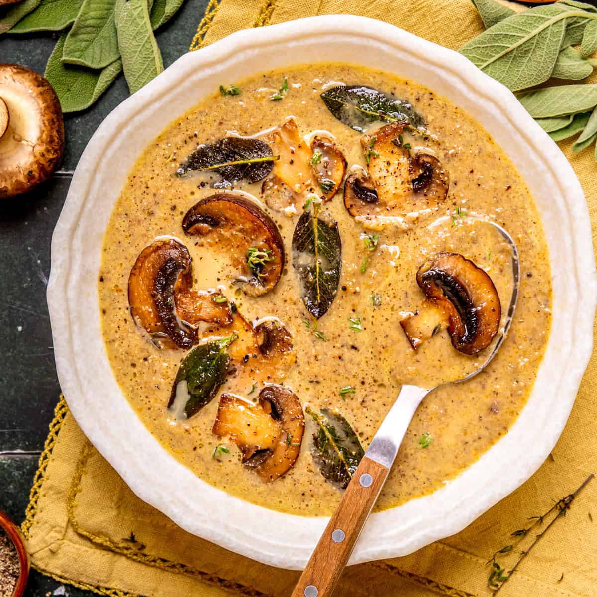 A bowl of creamy mushroom soup garnished with sautéed mushrooms and fresh sage leaves, with a spoon resting inside.