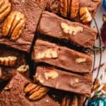 Close-up of chocolate fudge squares topped and filled with pecan halves, stacked on a wire rack.