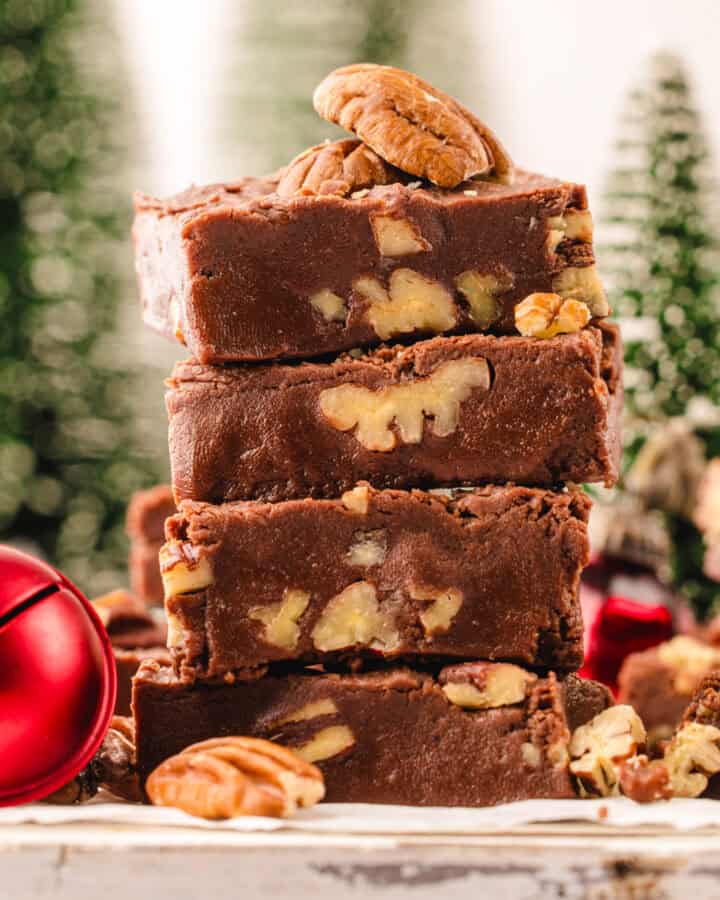 Four pieces of chocolate fudge with nuts are stacked on a wooden surface, garnished with pecans.