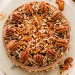 A round chocolate cake topped with chopped pecans, shredded coconut, chocolate frosting swirls, and whole pecans, displayed on a white surface.
