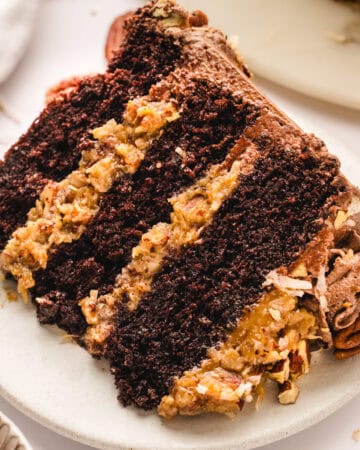 A close-up of a slice of German chocolate cake with three layers of chocolate cake and coconut-pecan filling, served on a white plate.