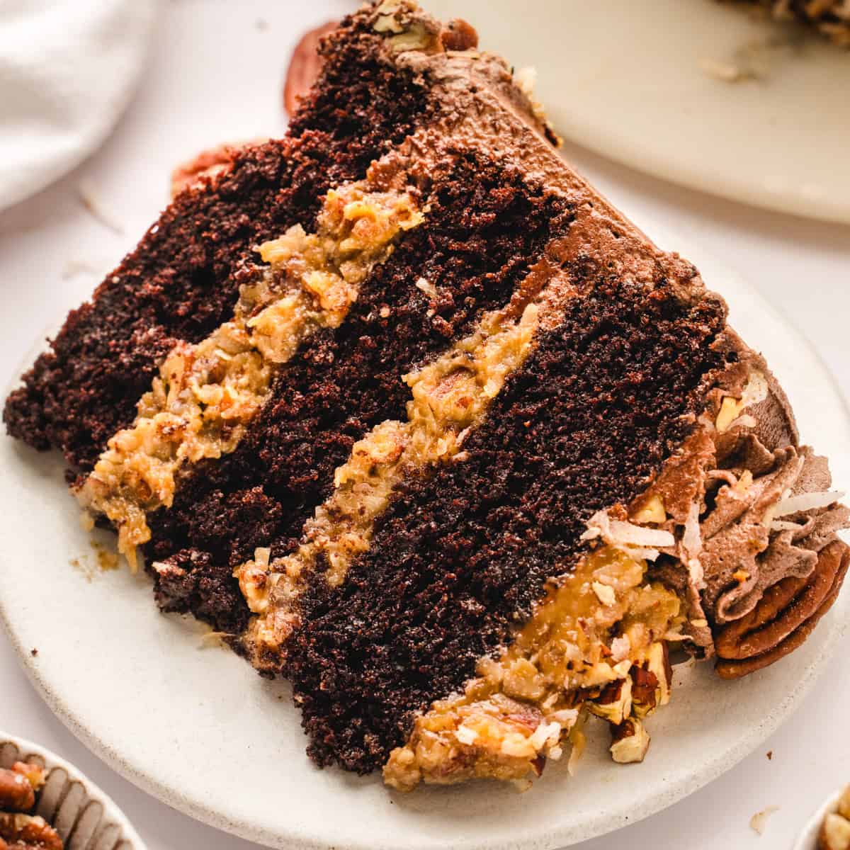 A close-up of a slice of German chocolate cake with three layers of chocolate cake and coconut-pecan filling, served on a white plate.