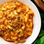 A white bowl filled with creamy macaroni and cheese topped with ground beef and sprinkled with fresh herbs and black pepper.