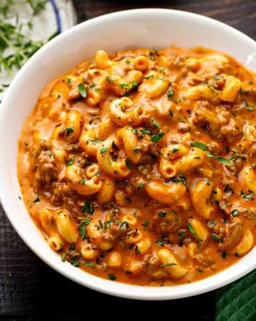 A white bowl filled with creamy macaroni pasta mixed with ground meat and garnished with chopped herbs.
