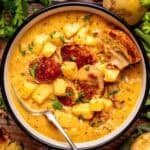 A bowl of creamy soup with sausage slices, potato chunks, herbs, and a piece of toasted bread, garnished with chopped parsley and black pepper.