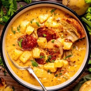 A bowl of creamy soup with sausage slices, potato chunks, herbs, and a piece of toasted bread, garnished with chopped parsley and black pepper.