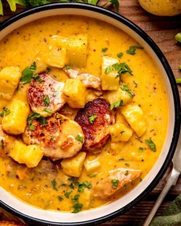 A bowl of creamy potato and sausage soup garnished with parsley sits on a wooden table next to a spoon, celery, and bread slices.