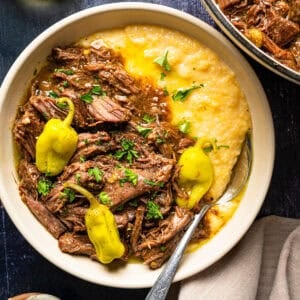 A bowl of shredded beef pot roast served over creamy grits, garnished with parsley and yellow peppers.