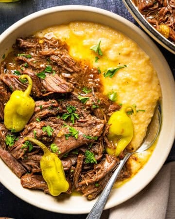 A bowl of creamy grits topped with shredded beef roast, yellow pepperoncini peppers, gravy, and fresh parsley, served with a spoon.