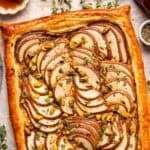 Rectangular pear tart with golden puff pastry crust, topped with thinly sliced pears, pistachios, and fresh herbs, displayed on parchment paper.