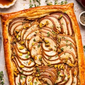 Rectangular pear tart with golden puff pastry crust, topped with thinly sliced pears, pistachios, and fresh herbs, displayed on parchment paper.