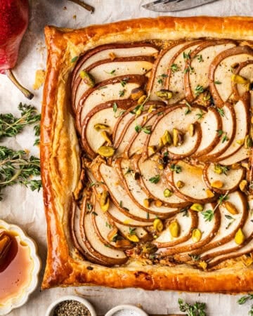 A rectangular pear tart with golden puff pastry, topped with thinly sliced pears, chopped nuts, and fresh thyme.