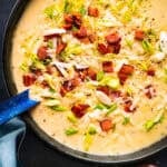 Creamy potato soup topped with crispy bacon, shredded cheese, chopped green onions, and cracked black pepper in a black pot with a blue ladle.