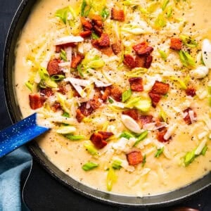 Creamy potato soup topped with crispy bacon, shredded cheese, chopped green onions, and cracked black pepper in a black pot with a blue ladle.