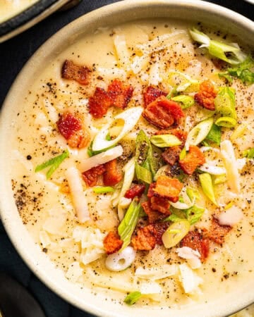 A creamy soup topped with crispy bacon pieces, sliced green onions, shredded cheese, and cracked black pepper, served in a beige bowl on a dark surface.