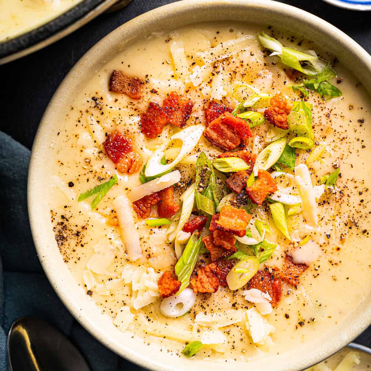 A creamy soup topped with crispy bacon pieces, sliced green onions, shredded cheese, and cracked black pepper, served in a beige bowl on a dark surface.