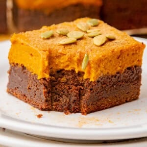 A square piece of dessert with a chocolate base and thick orange pumpkin topping, garnished with pumpkin seeds, sits on a white plate.