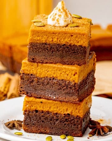 A stack of three pumpkin and chocolate layered bars topped with whipped cream and sprinkled with pumpkin seeds.