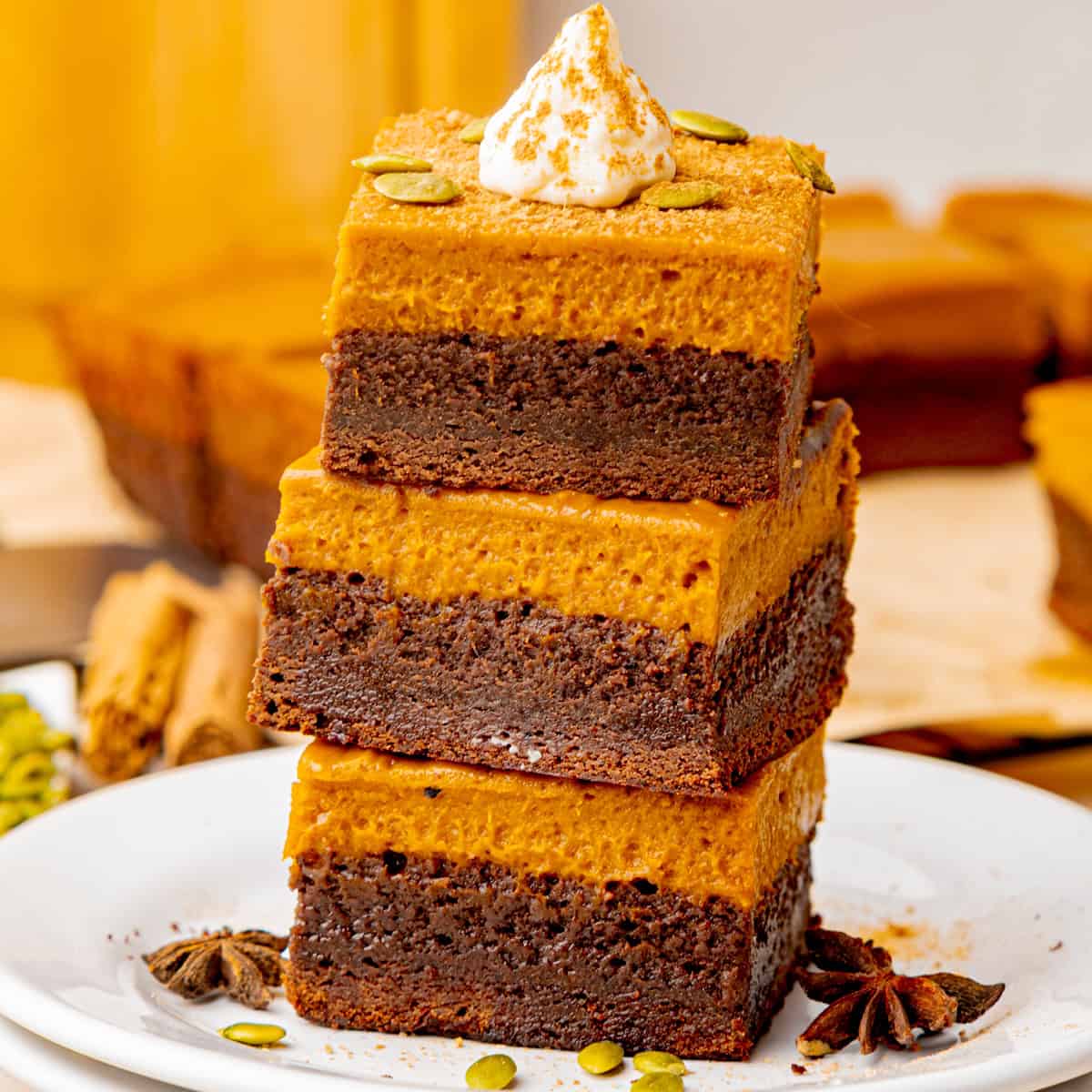 A stack of three pumpkin and chocolate layered bars topped with whipped cream and sprinkled with pumpkin seeds.