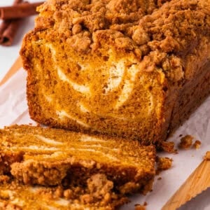 A loaf of pumpkin bread with a cream cheese swirl and crumb topping sits on parchment paper, with one slice cut and lying in front of the loaf.