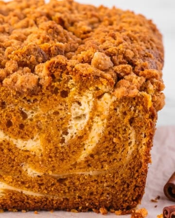 A close-up of a sliced loaf of pumpkin bread with a cream cheese swirl and crumb topping, sitting on parchment paper.