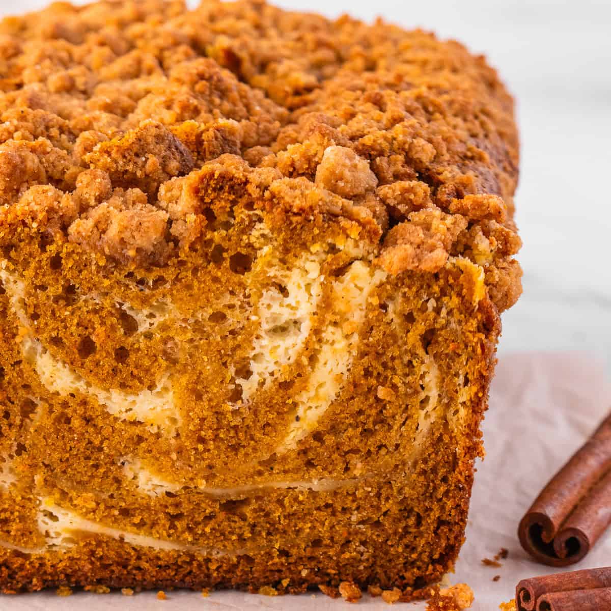 A close-up of a sliced loaf of pumpkin bread with a cream cheese swirl and crumb topping, sitting on parchment paper.