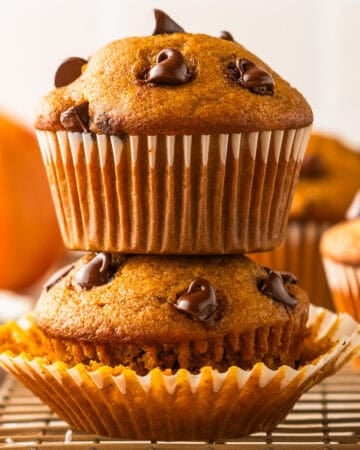 Two chocolate chip muffins are stacked on a cooling rack. The top muffin is upright, while the bottom muffin is on its side with the wrapper partially opened.