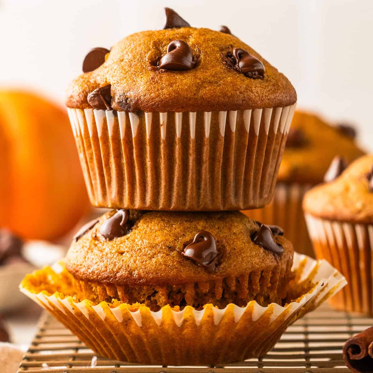 Two chocolate chip muffins are stacked on a cooling rack. The top muffin is upright, while the bottom muffin is on its side with the wrapper partially opened.