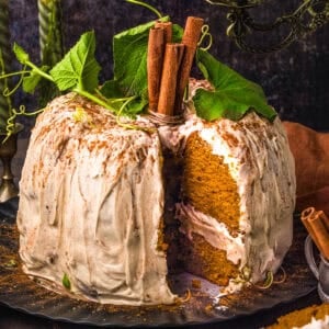 Pumpkin spice cake with white frosting, topped with cinnamon sticks and green leaves, sits on a black plate. A large slice has been cut, revealing the moist interior.