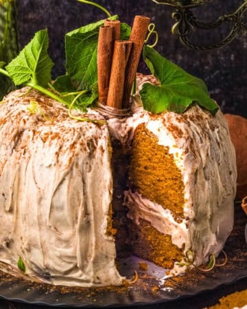 A frosted pumpkin cake with a slice missing, topped with cinnamon sticks and green leaves, sits on a black plate.