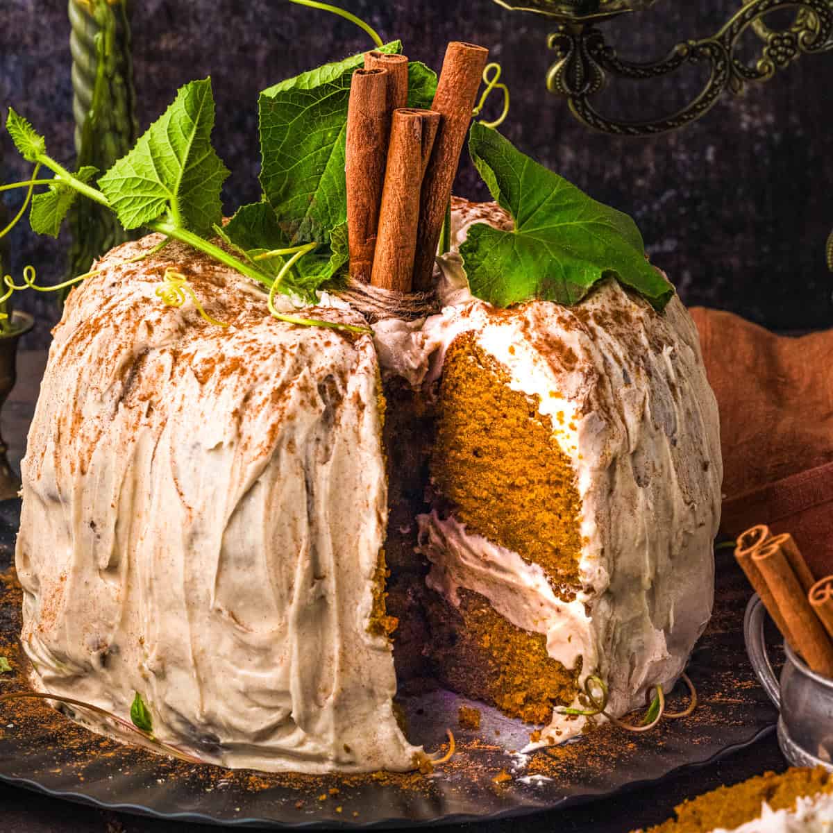 A frosted pumpkin cake with a slice missing, topped with cinnamon sticks and green leaves, sits on a black plate.
