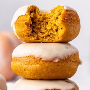 Three frosted pumpkin donuts are stacked. The top donut has a large bite taken out, revealing a moist, orange interior.