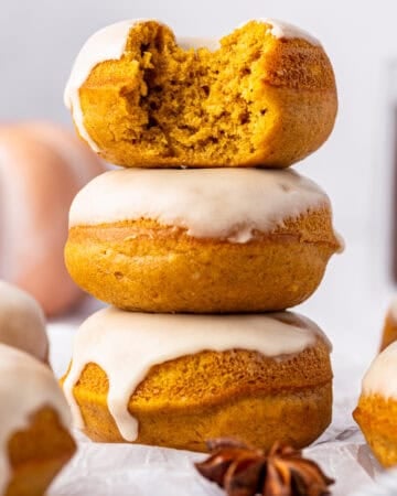Three pumpkin donuts with white glaze stacked, the top one has a bite taken out. Other donuts and a pumpkin are blurred in the background.