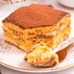 A slice of pumpkin tiramisu dusted with cocoa powder on a white plate, with a fork holding a bite-sized piece in the foreground.