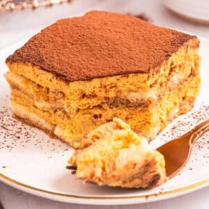 A slice of pumpkin tiramisu dusted with cocoa powder on a white plate, with a fork holding a bite-sized piece in the foreground.