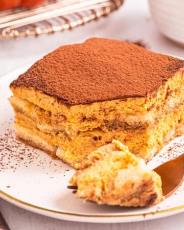 A slice of tiramisu dusted with cocoa powder on a white plate, with a fork holding a bite in the foreground.