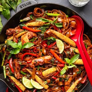 A skillet filled with stir-fried noodles, chicken, baby corn, red peppers, basil, lime wedges, and sliced chili, garnished with fresh herbs, with red serving utensils beside the dish.
